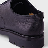 A close-up rear three-quarter view of a dark purple leather lace-up shoe with a chunky black lug sole, shown on a light gray background.