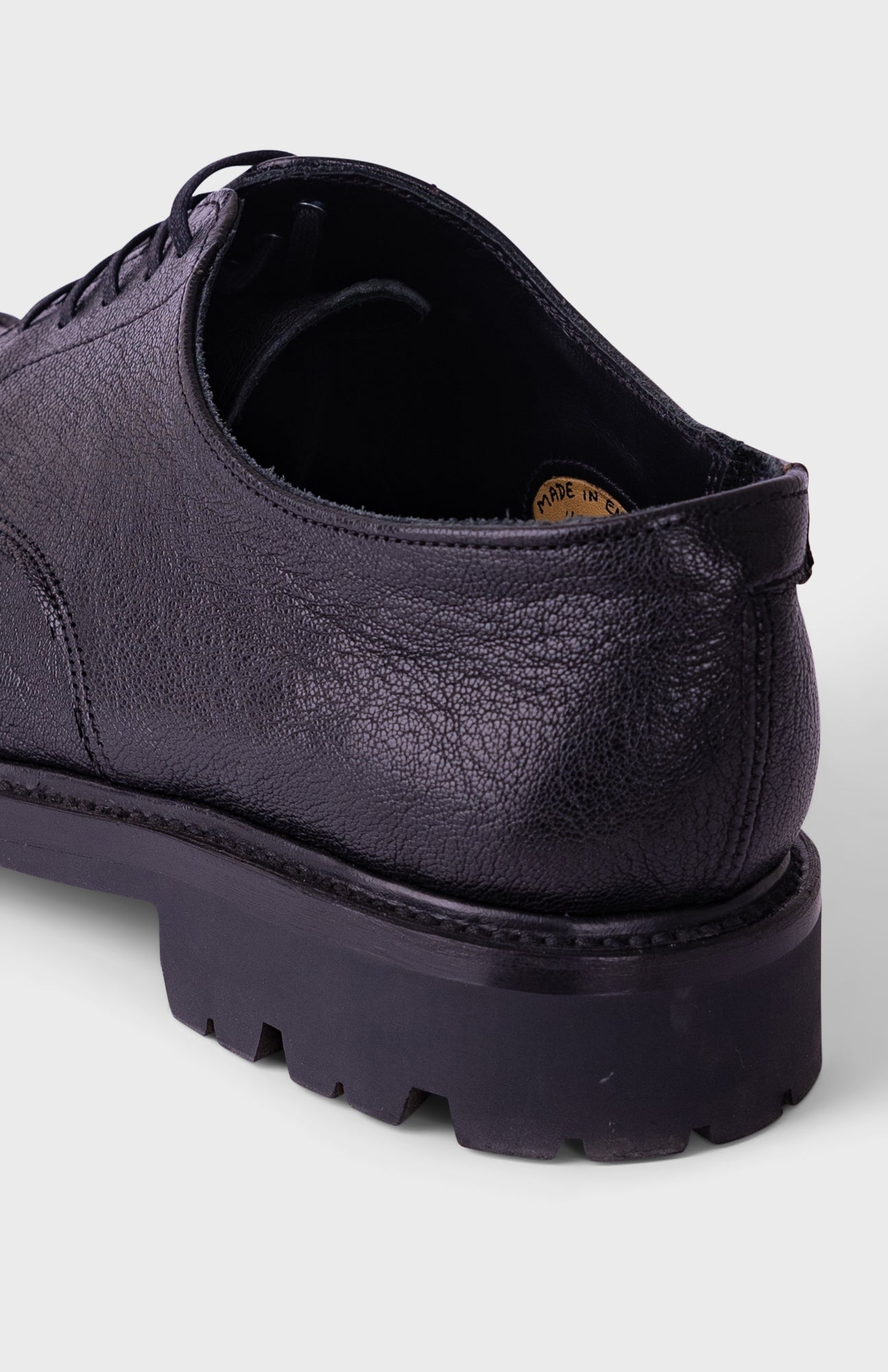 A close-up rear three-quarter view of a dark purple leather lace-up shoe with a chunky black lug sole, shown on a light gray background.