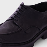 A close-up shot of a black leather lace-up shoe with a chunky lug sole, shown against a light gray background.