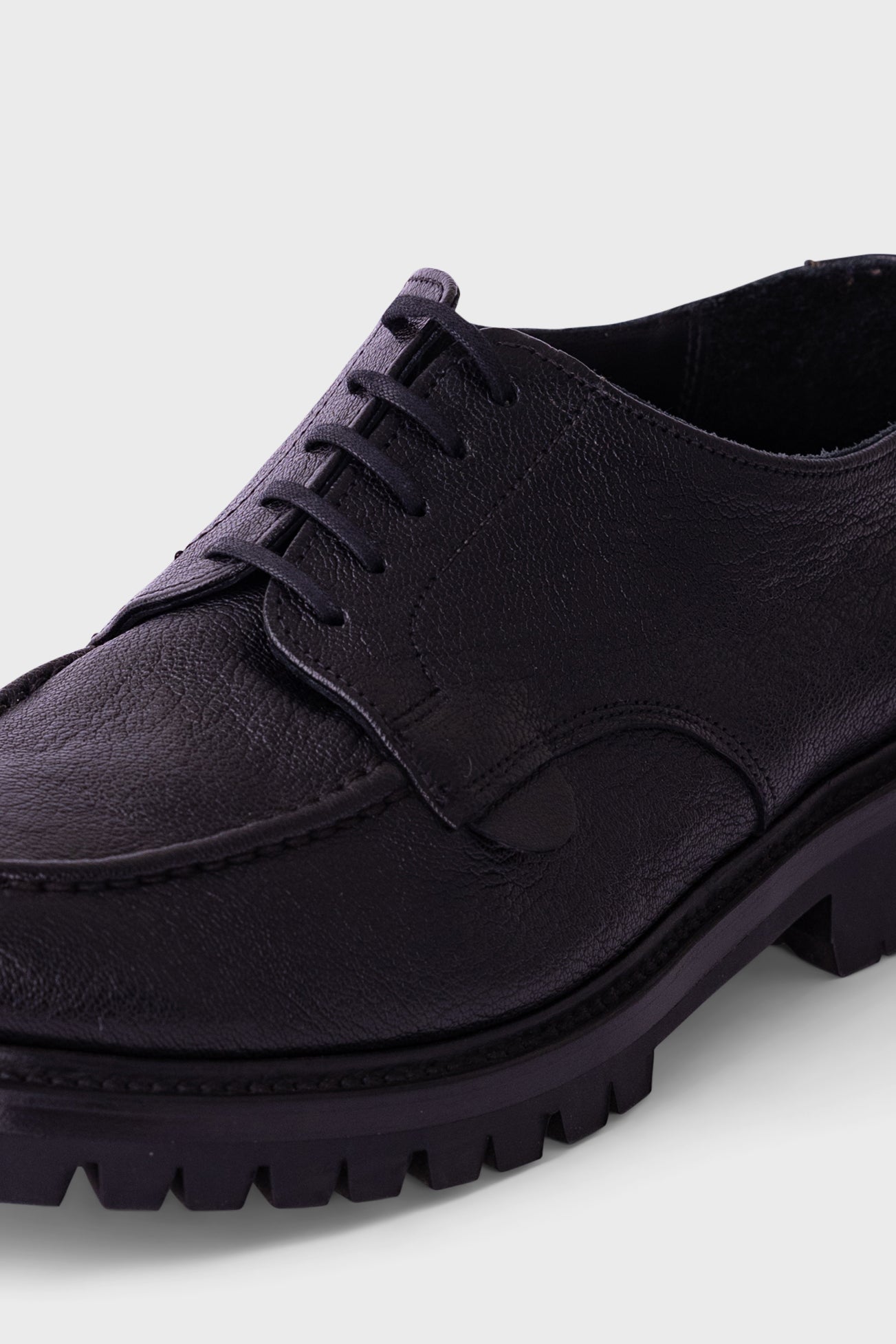 A close-up shot of a black leather lace-up shoe with a chunky lug sole, shown against a light gray background.