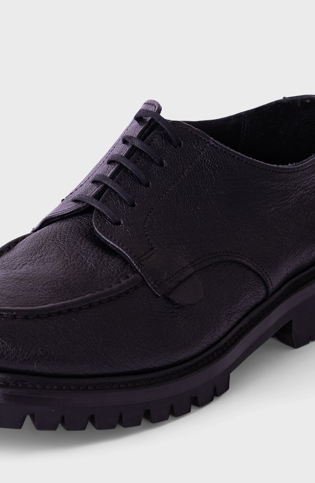 A close-up shot of a black leather lace-up shoe with a chunky lug sole, shown against a light gray background.