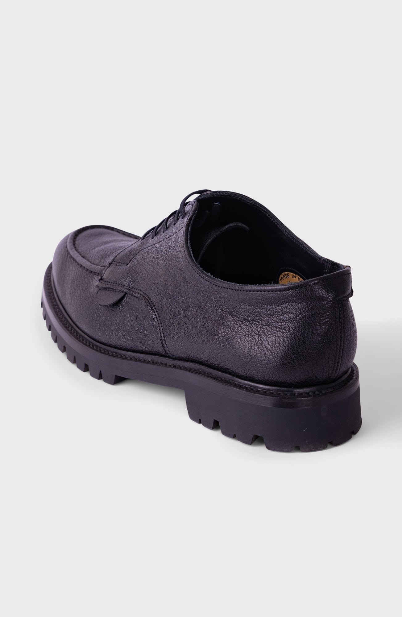A single black leather lace-up shoe with a chunky lug sole, shown from a rear three-quarter angle on a light gray background.