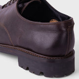 A close-up shot of a dark brown leather lace-up shoe, showing the side profile with a chunky lug sole, on a light gray background.