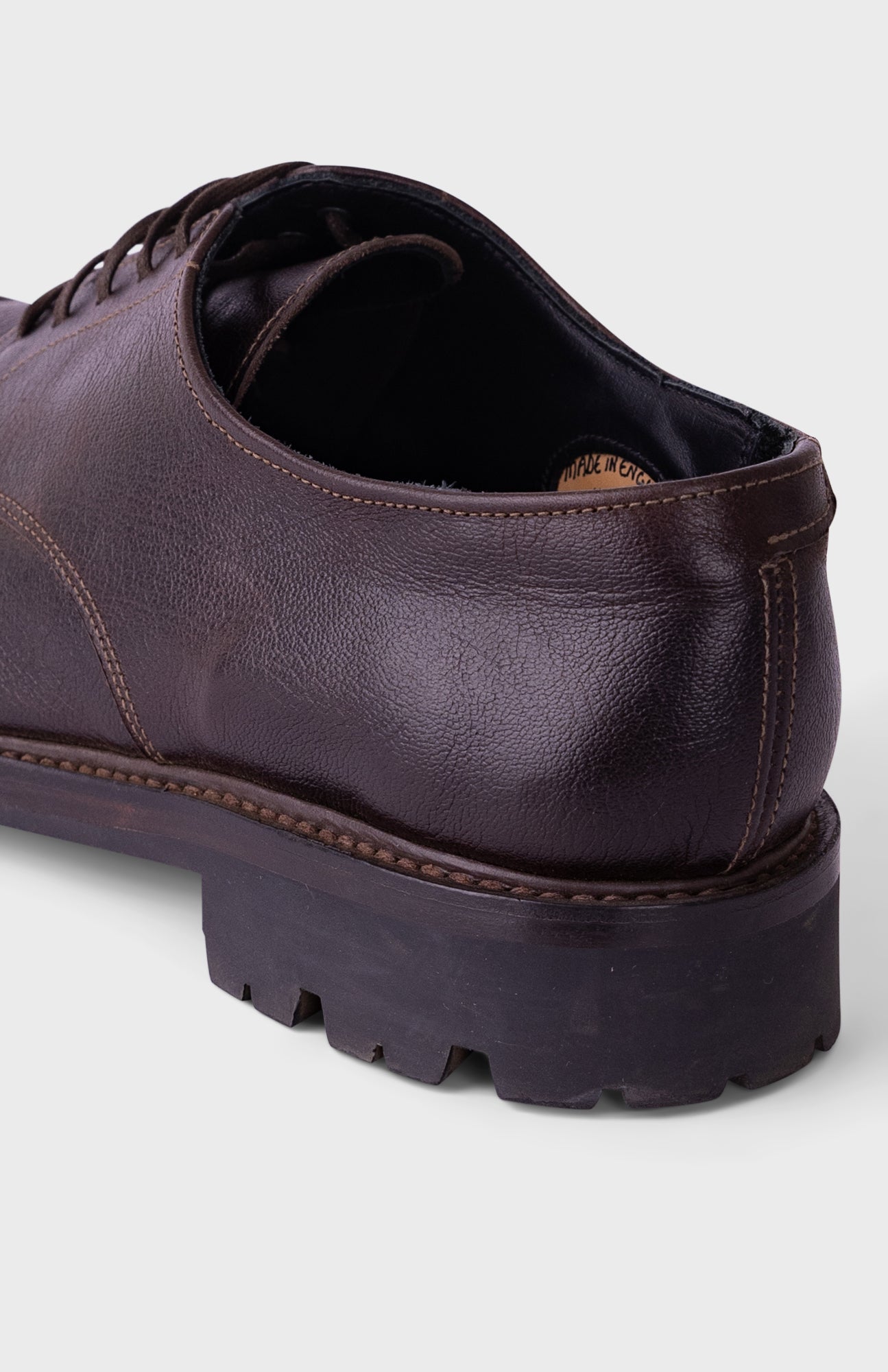 A close-up shot of a dark brown leather lace-up shoe, showing the side profile with a chunky lug sole, on a light gray background.