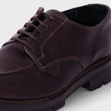 A close-up shot of a dark brown leather lace-up shoe with a rugged, chunky black sole, shown against a light gray background.
