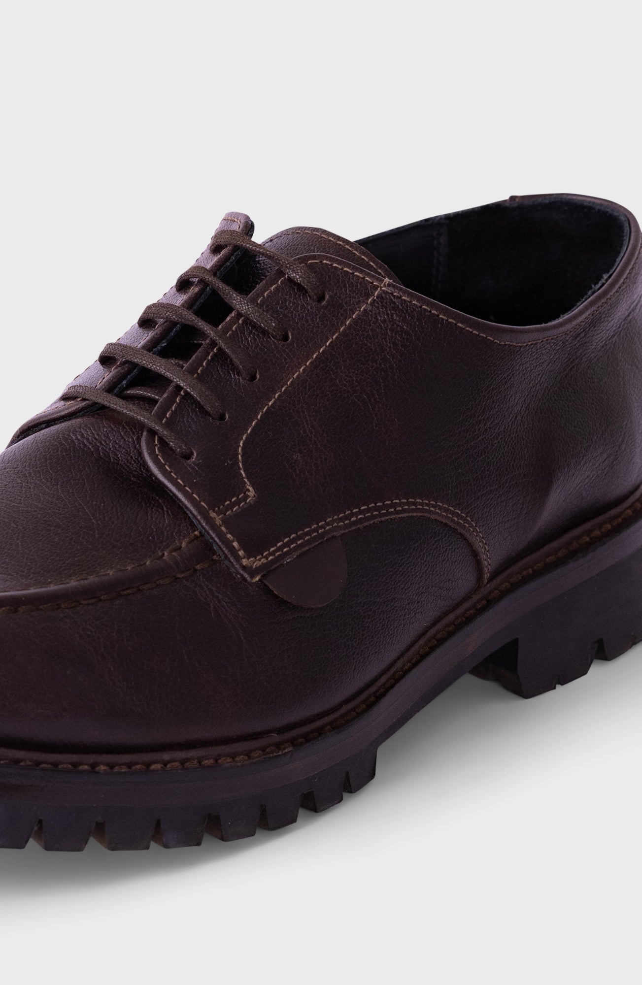 A close-up shot of a dark brown leather lace-up shoe with a rugged, chunky black sole, shown against a light gray background.