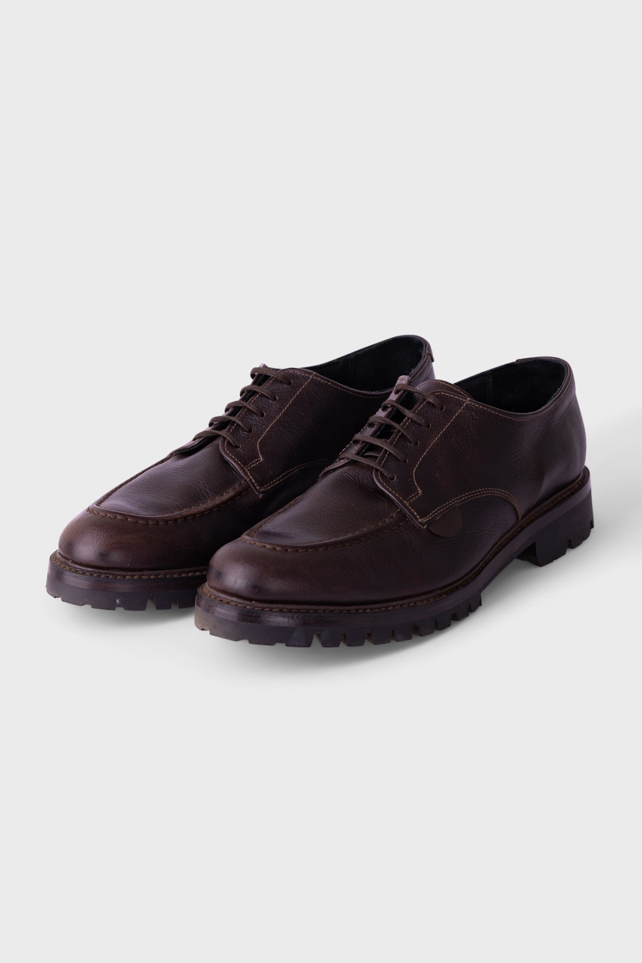 A pair of dark brown leather lace-up shoes with a rugged lug sole, displayed on a light gray background.