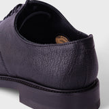 A close-up shot of a dark purple textured leather derby shoe, showing the side profile, laces, and thick black sole against a light gray background.