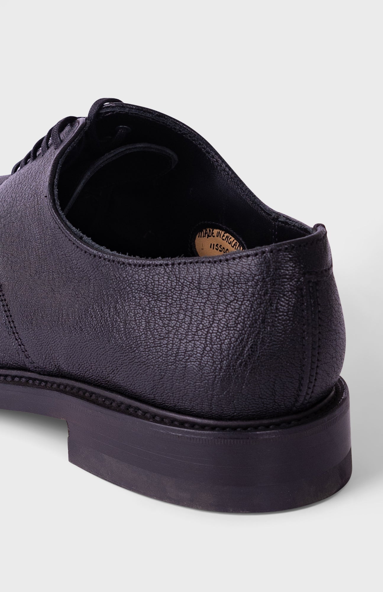A close-up shot of a dark purple textured leather derby shoe, showing the side profile, laces, and thick black sole against a light gray background.