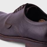 A close-up shot of a dark brown leather derby shoe, showing the textured leather upper, laces, and stacked rubber sole against a light gray background.