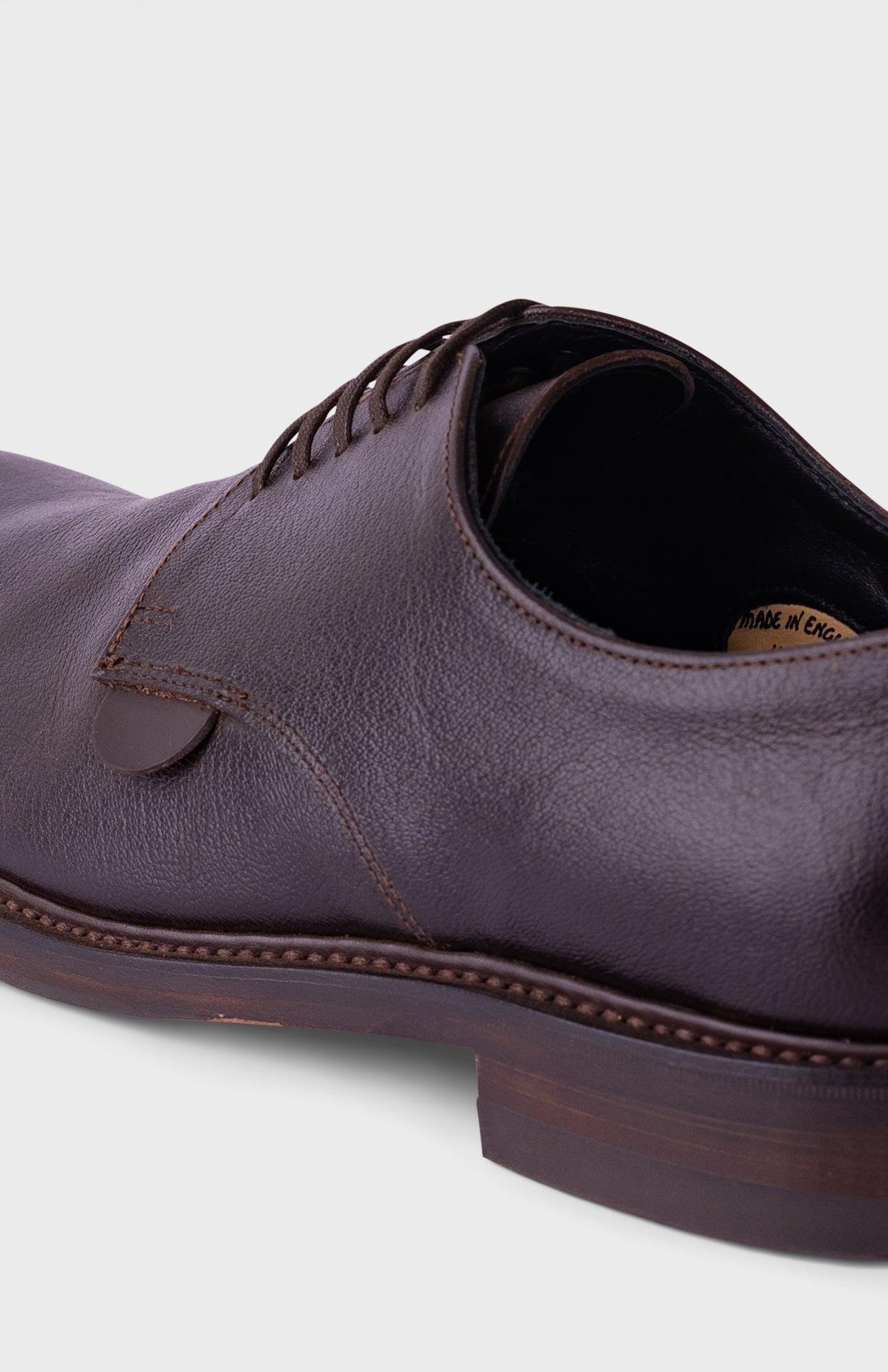 A close-up shot of a dark brown leather derby shoe, showing the textured leather upper, laces, and stacked rubber sole against a light gray background.