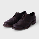 A pair of dark brown leather lace-up dress shoes with a low stacked heel, shown on a light gray background.