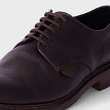A close-up shot of a dark brown leather oxford dress shoe with matching laces, showing the textured leather, stitching, and a low stacked heel against a light gray background.