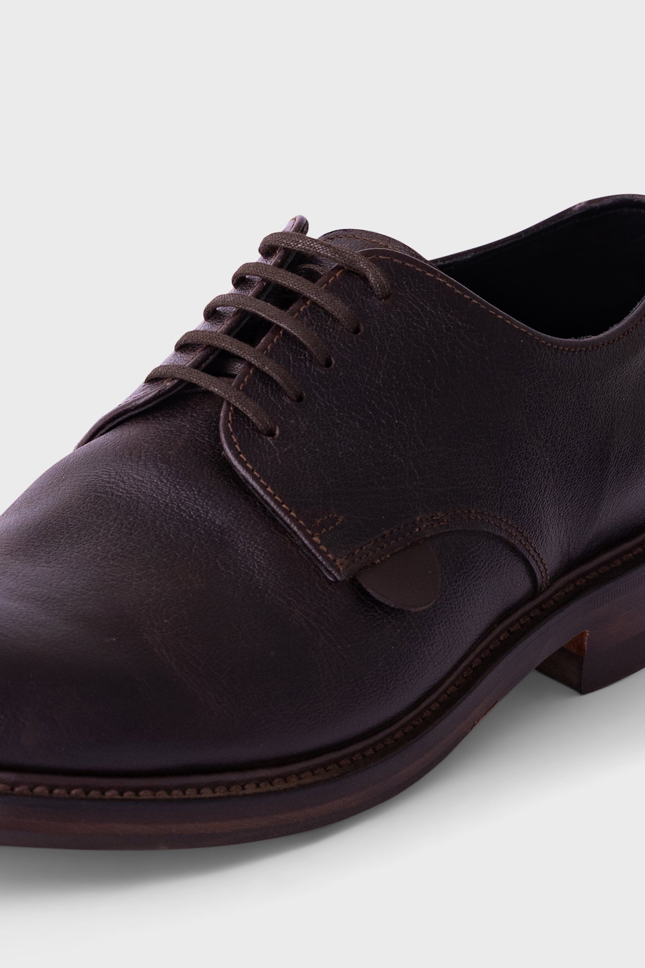 A close-up shot of a dark brown leather oxford dress shoe with matching laces, showing the textured leather, stitching, and a low stacked heel against a light gray background.