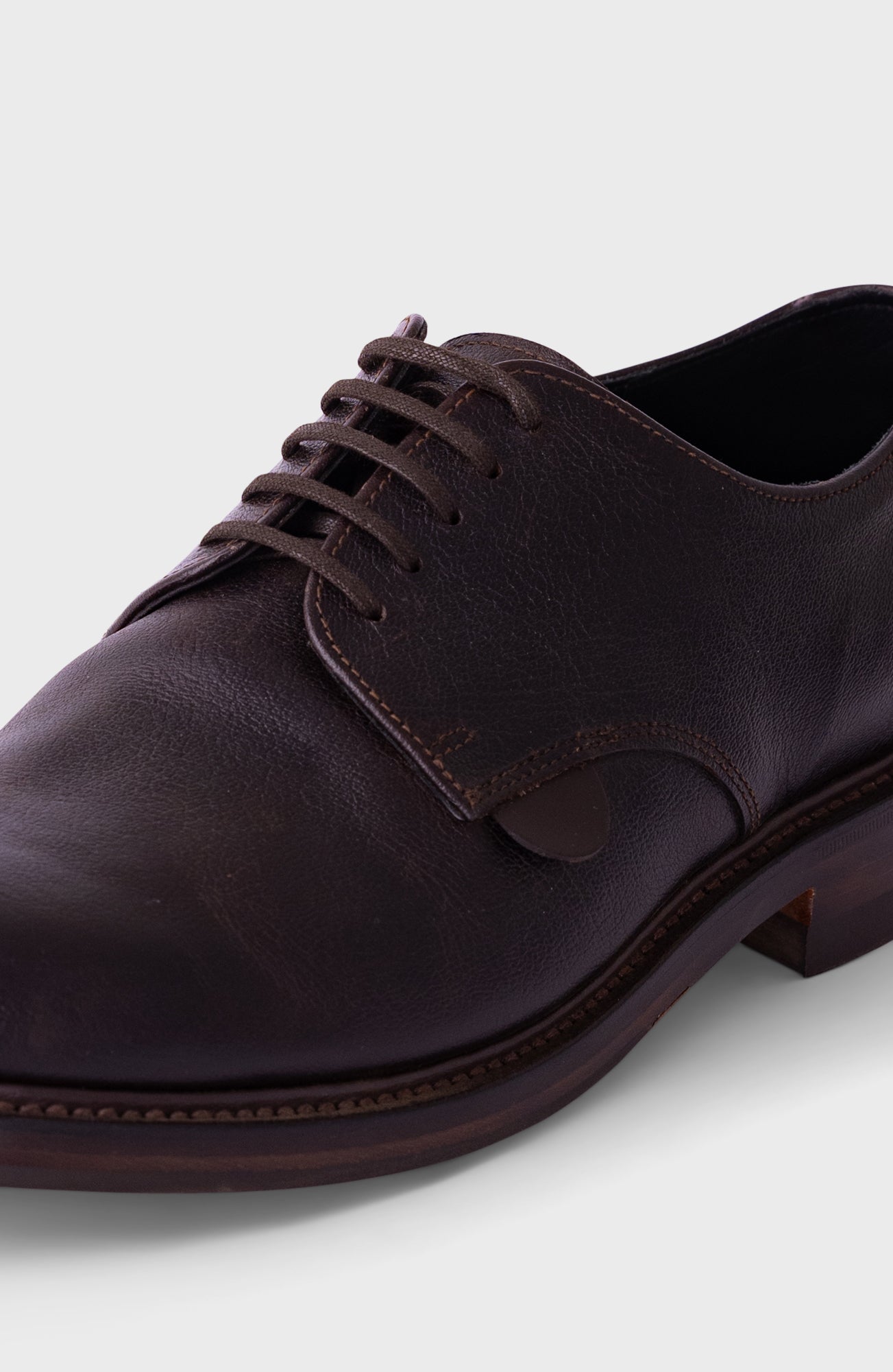 A close-up shot of a dark brown leather oxford dress shoe with matching laces, showing the textured leather, stitching, and a low stacked heel against a light gray background.