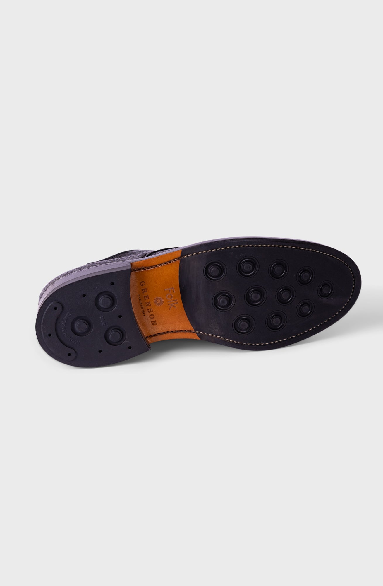 A close-up of a shoe sole showing a dark navy or black rubber outsole with circular tread patterns, a tan leather insole, and a dark purple side edge, photographed on a light gray background.