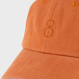 A close-up shot of a washed orange baseball cap with a tonal embroidered double-ring logo on the front, set against a light gray background.