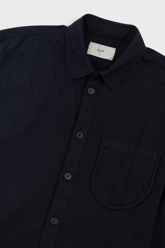 Close-up of a dark navy button-up shirt with a collar, chest pocket, and a visible Folk label inside the neck, shot against a light gray background.