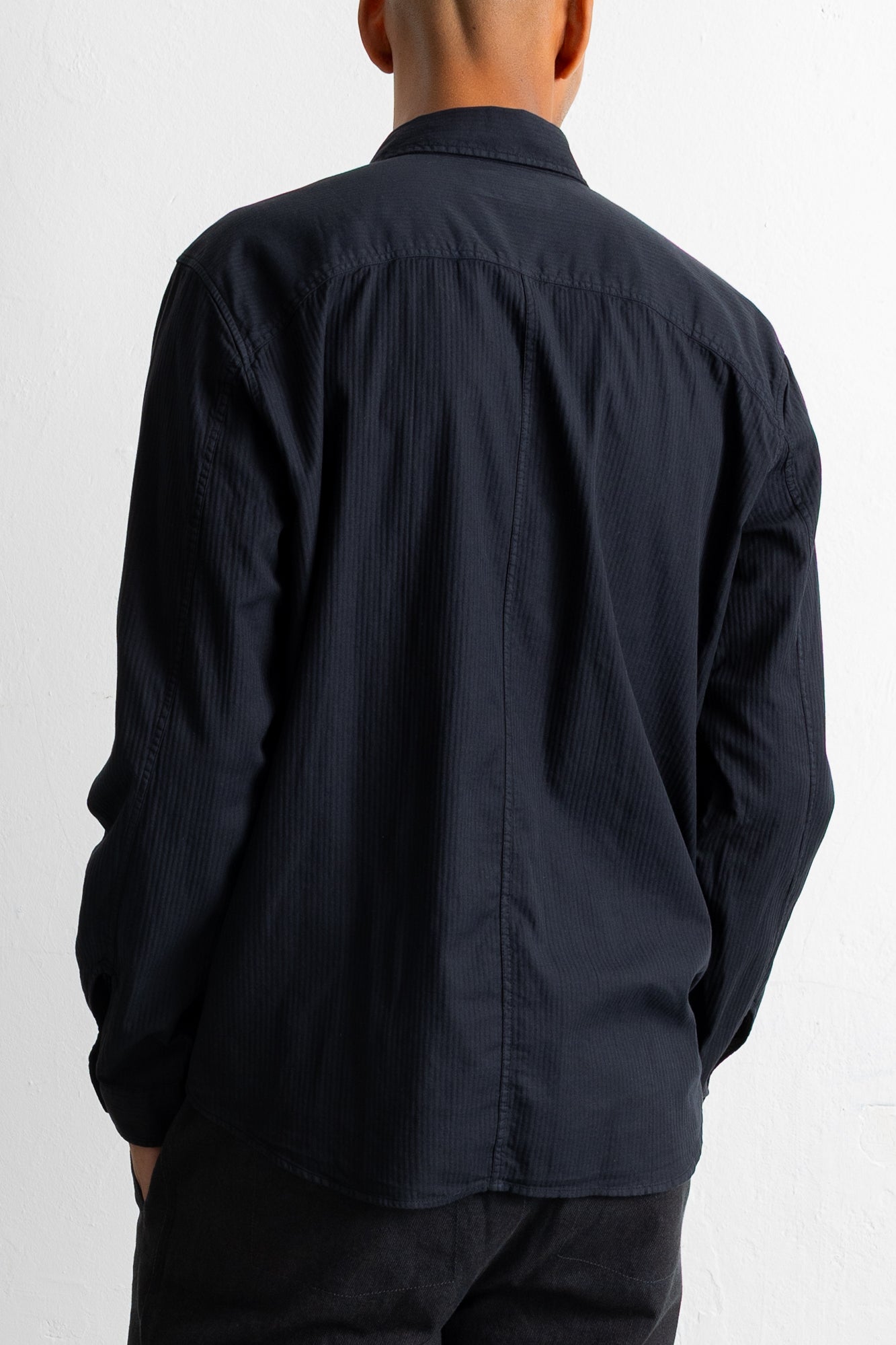 A man viewed from behind wearing a black long-sleeve button-down shirt with a subtle vertical textured stripe pattern.