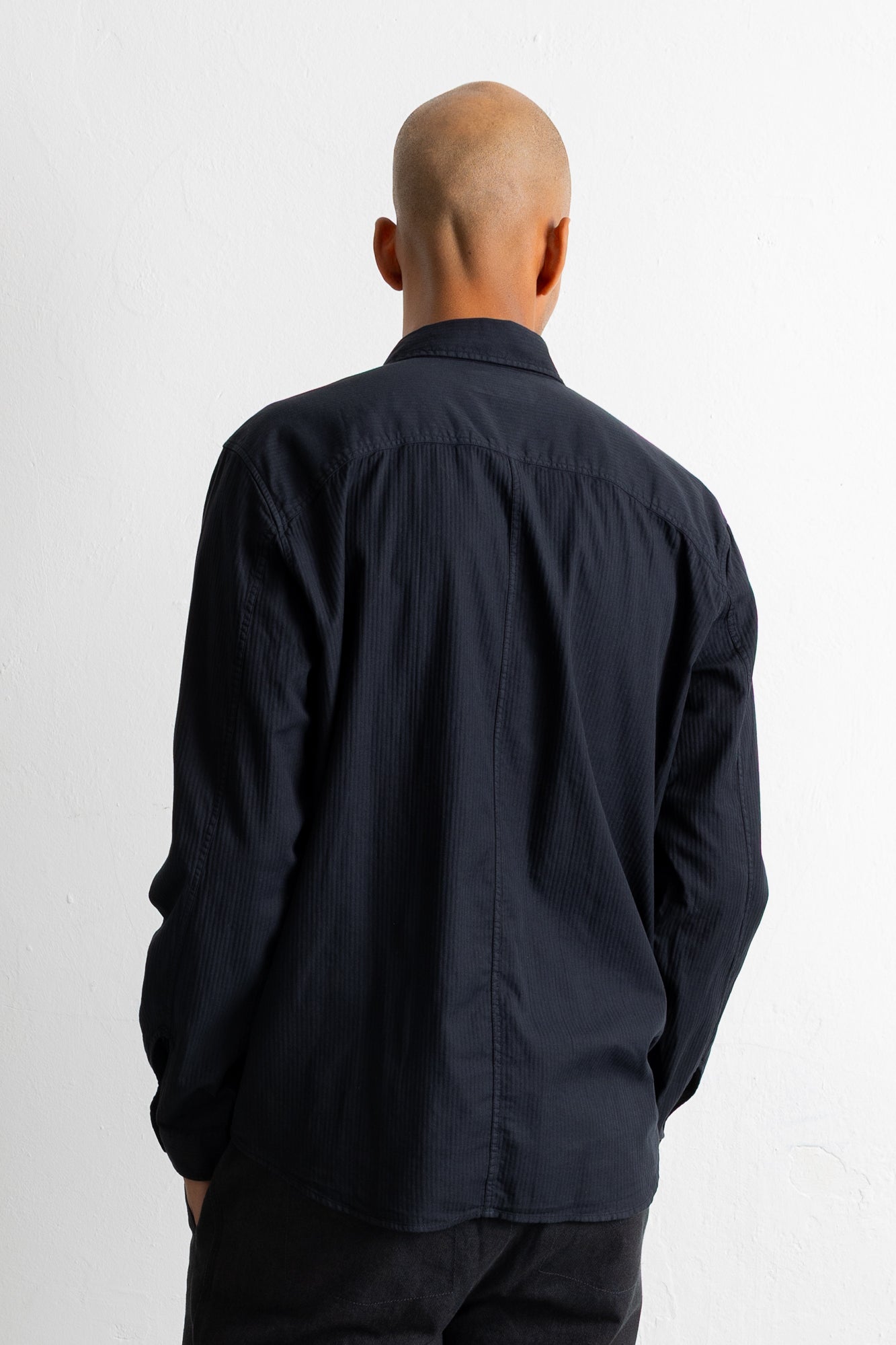 A man viewed from behind wearing a black long-sleeve button-up shirt and dark pants, standing against a plain white background.