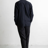 A man viewed from behind wearing a black long-sleeve button-up shirt, dark charcoal pants, and black sneakers with white soles, standing against a plain white wall.