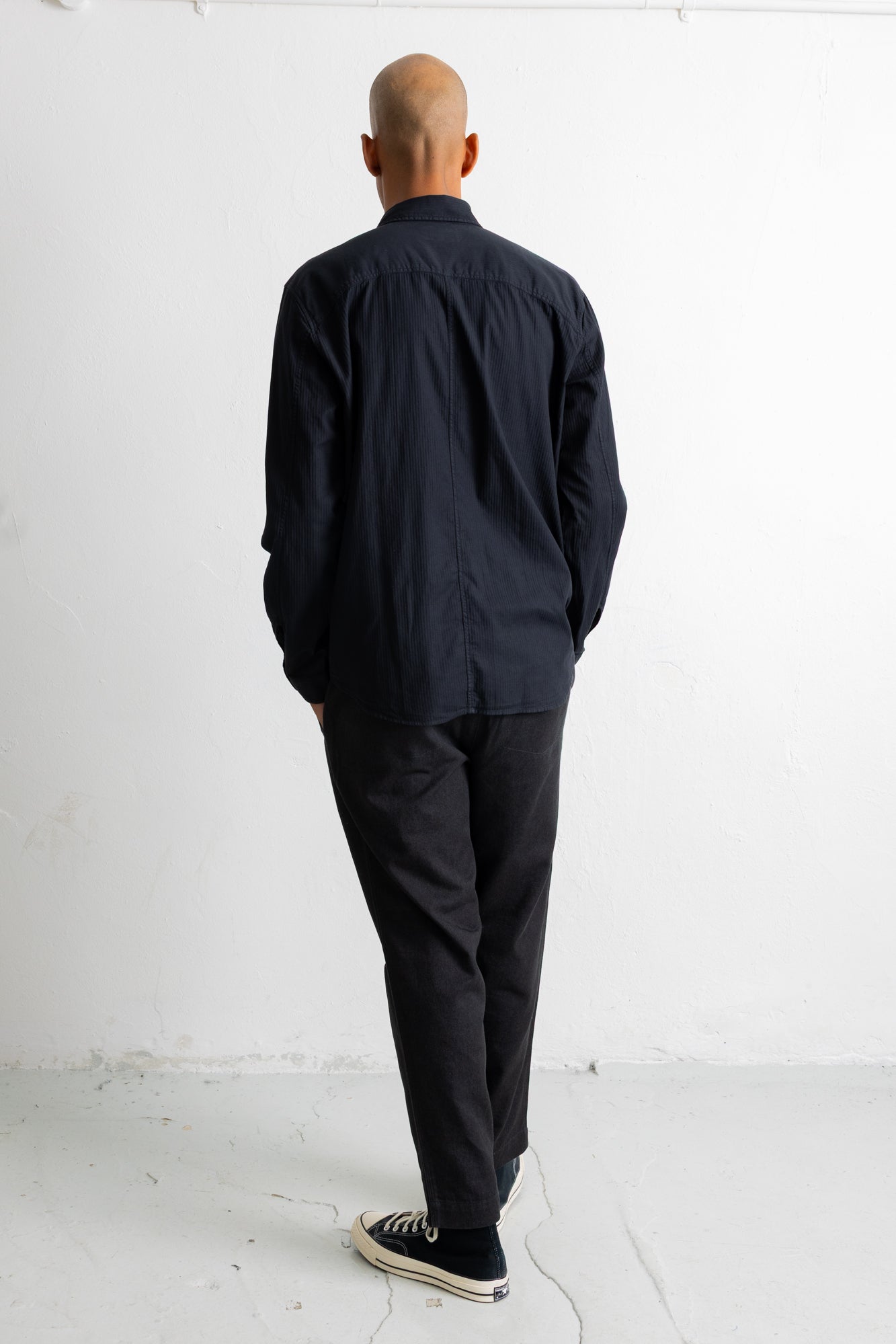A man viewed from behind wearing a black long-sleeve button-up shirt, dark charcoal pants, and black sneakers with white soles, standing against a plain white wall.