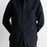A man wearing a black long-sleeve button-down shirt with a chest pocket, paired with dark trousers, photographed against a light off-white wall.