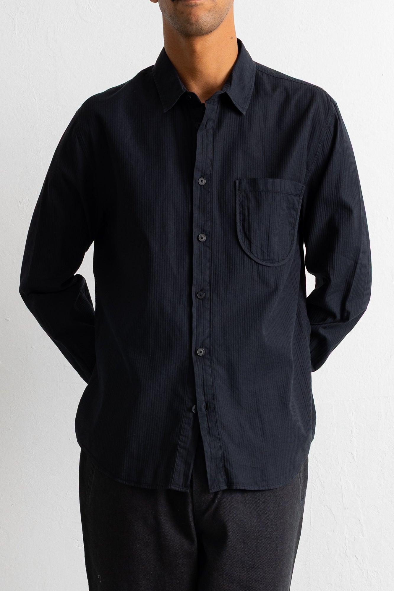 A man wearing a black long-sleeve button-down shirt with a chest pocket, paired with dark trousers, photographed against a light off-white wall.