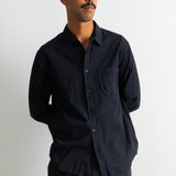 A man wearing a black long-sleeve button-down shirt with a chest pocket, paired with dark trousers, standing against a plain white wall.