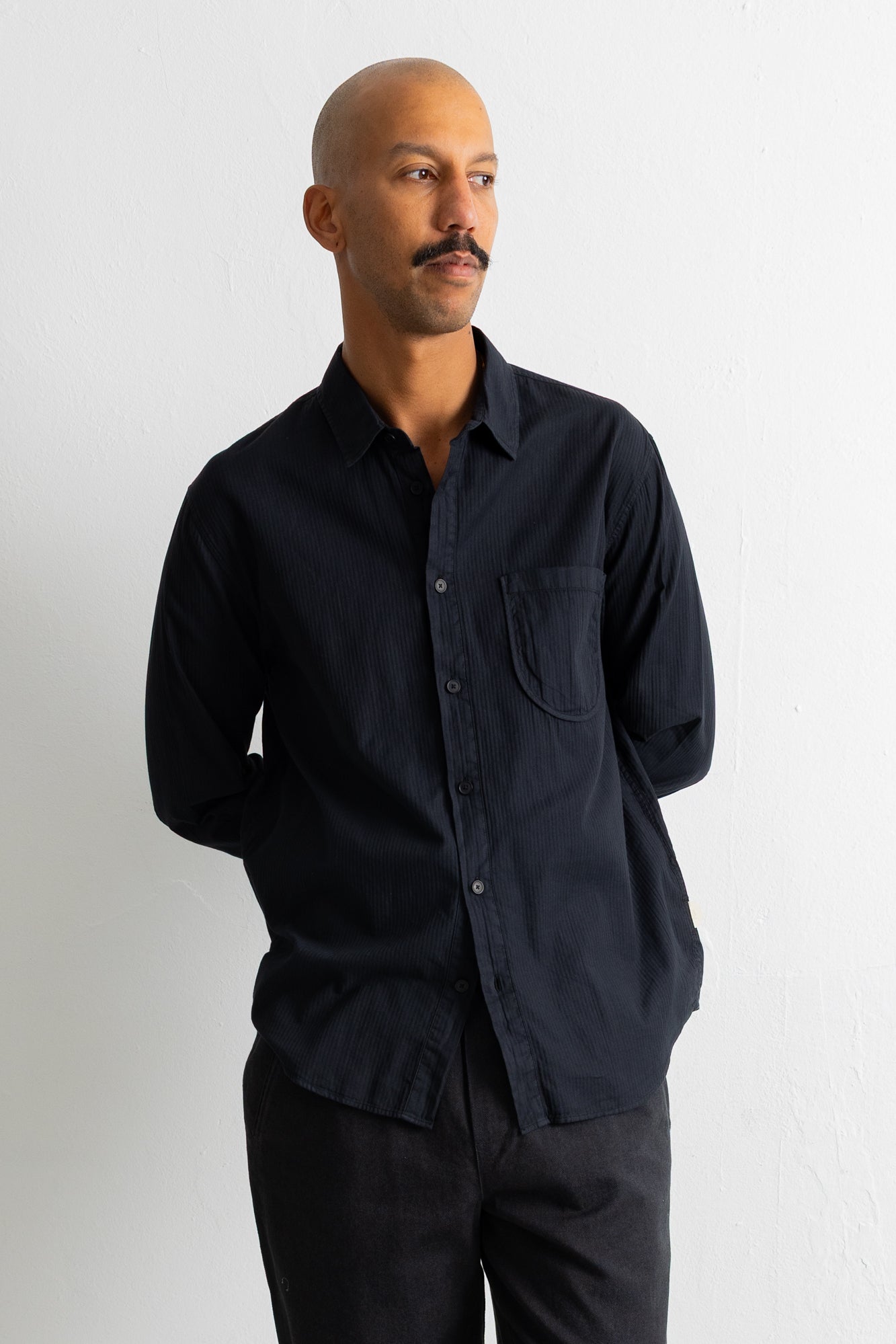 A man wearing a black long-sleeve button-down shirt with a chest pocket, paired with dark trousers, standing against a plain white wall.