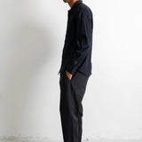 A man in a black long-sleeve button-up shirt, dark charcoal trousers, and navy sneakers with white laces stands in profile against a plain white wall.