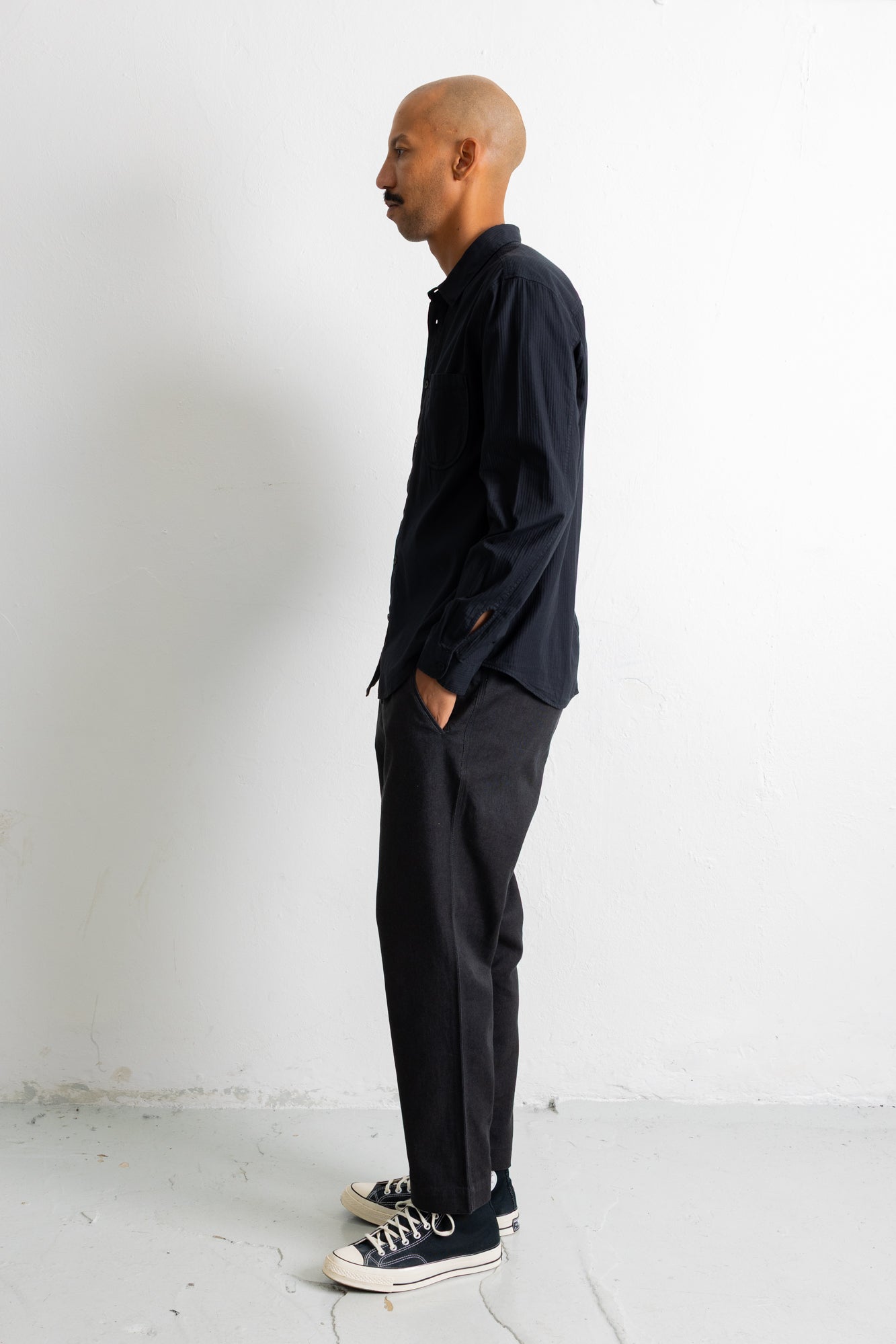 A man in a black long-sleeve button-up shirt, dark charcoal trousers, and navy sneakers with white laces stands in profile against a plain white wall.