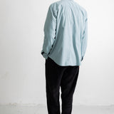 A man viewed from behind wearing a light blue long-sleeve button-up shirt, black pants, and black-and-white sneakers, standing against a plain white wall.