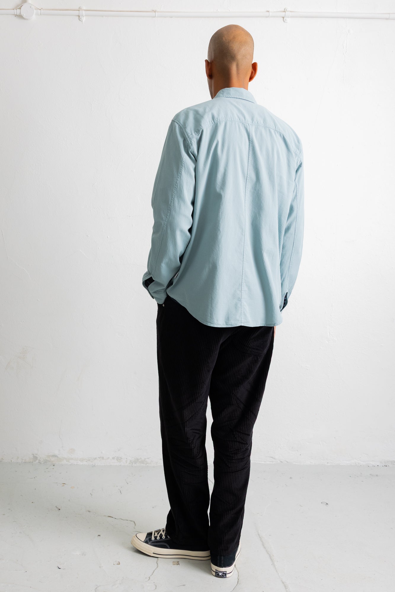 A man viewed from behind wearing a light blue long-sleeve button-up shirt, black pants, and black-and-white sneakers, standing against a plain white wall.