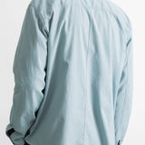 A man viewed from behind wearing a light blue pinstriped button-down shirt and dark trousers, set against a plain white studio background.