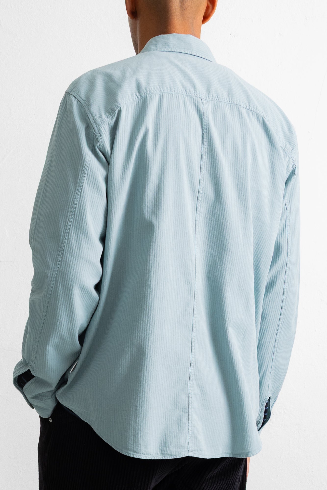 A man viewed from behind wearing a light blue pinstriped button-down shirt and dark trousers, set against a plain white studio background.