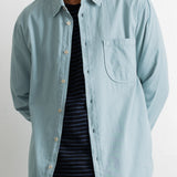 A man wearing a light blue pinstriped button-down shirt over a dark navy and white striped t-shirt, shown from the chin down against a plain white background.