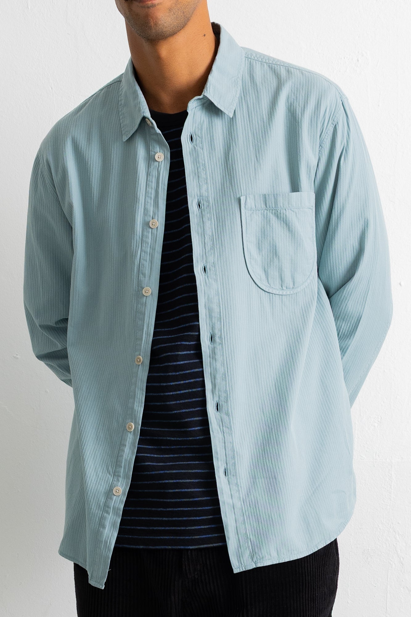 A man wearing a light blue pinstriped button-down shirt over a dark navy and white striped t-shirt, shown from the chin down against a plain white background.