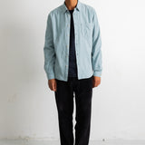 A man wearing a light blue button-down shirt over a dark striped t-shirt, black pants, and black-and-white sneakers, standing against a plain white wall.
