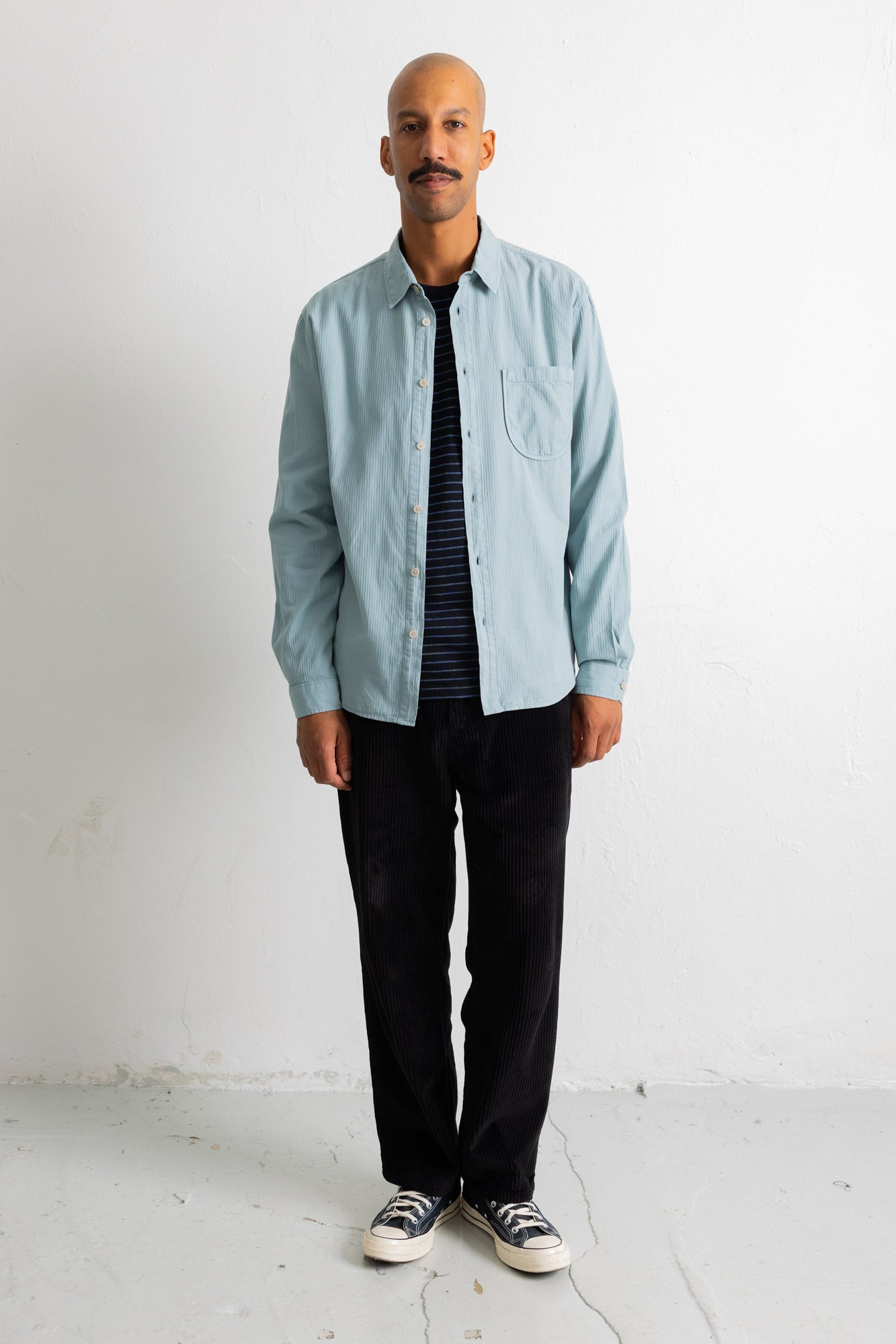 A man wearing a light blue button-down shirt over a dark striped t-shirt, black pants, and black-and-white sneakers, standing against a plain white wall.