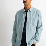A man wearing a light blue long-sleeve button-down shirt over a black and navy striped t-shirt, paired with black pants, standing against a plain white wall.