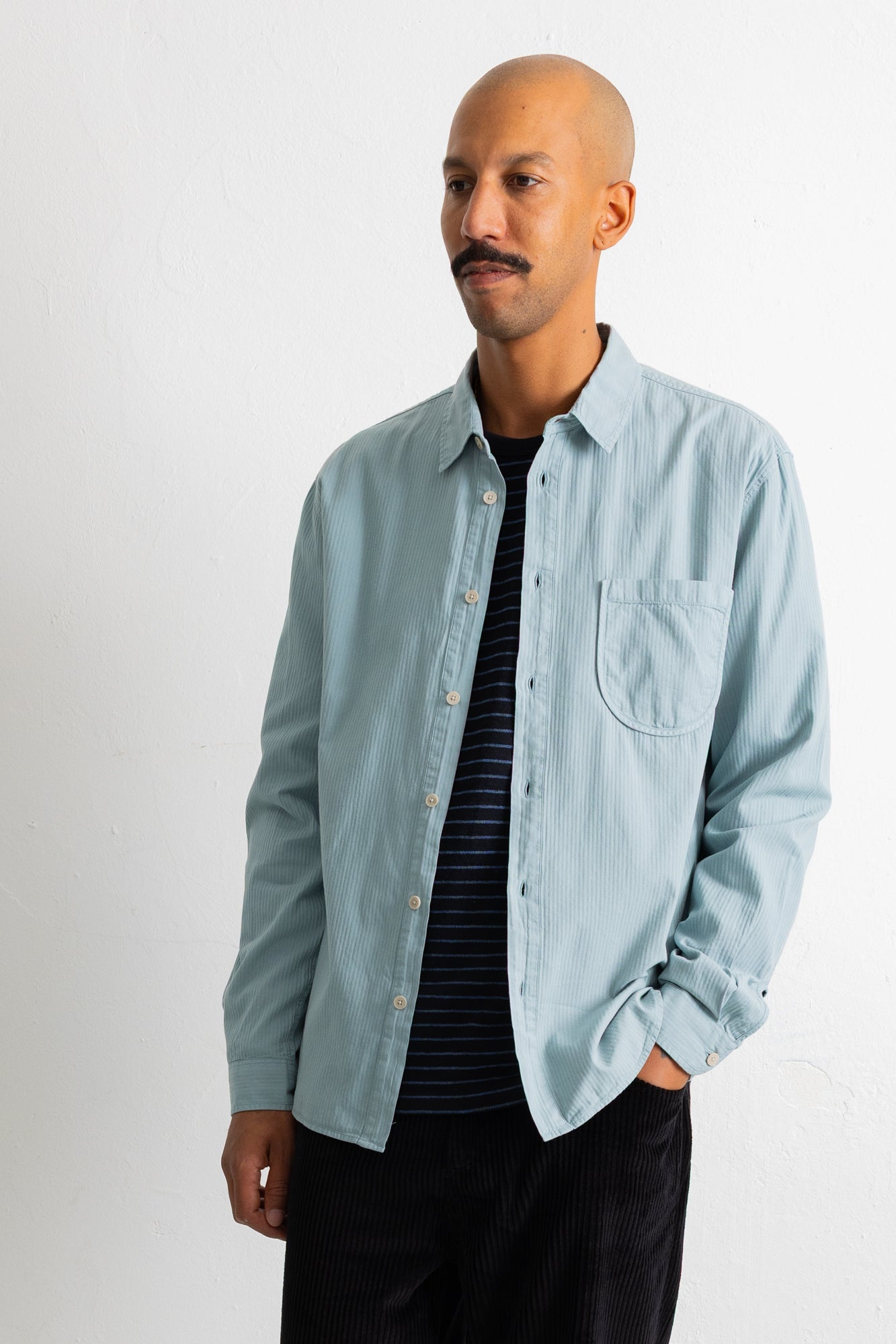 A man wearing a light blue long-sleeve button-down shirt over a black and navy striped t-shirt, paired with black pants, standing against a plain white wall.