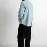 A man wearing a light blue long-sleeve button-up shirt, black pants, and black canvas sneakers, standing in profile against a white studio wall.