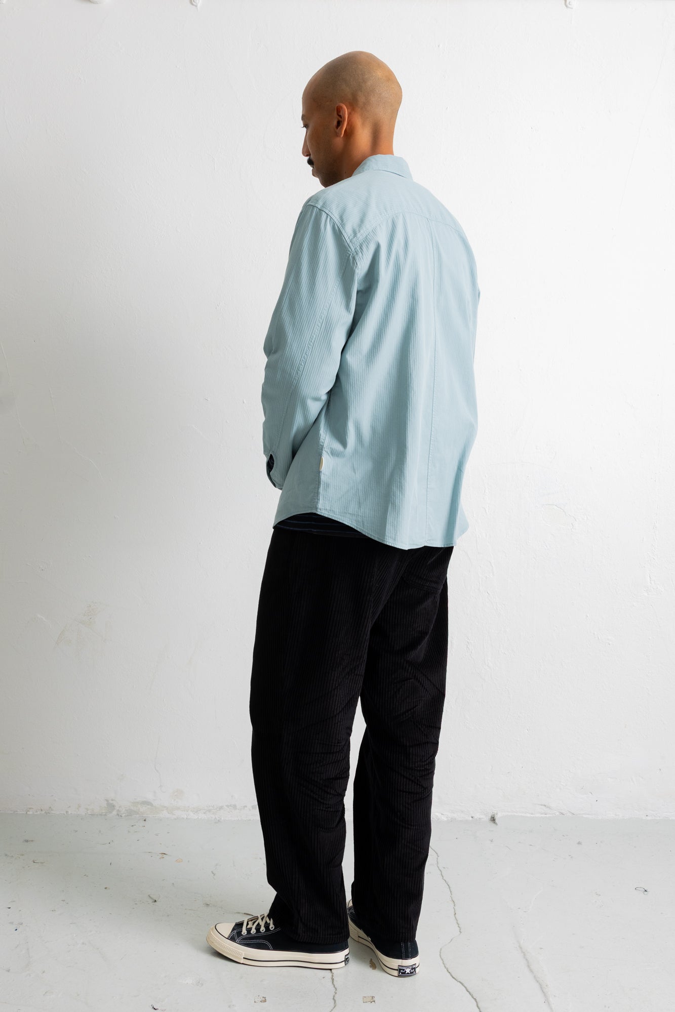 A man wearing a light blue long-sleeve button-up shirt, black pants, and black canvas sneakers, standing in profile against a white studio wall.