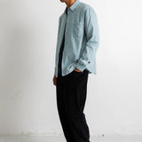 A man wearing a light blue long-sleeve button-down shirt, a black and white striped tie, black trousers, and black canvas sneakers, standing in profile against a white wall.