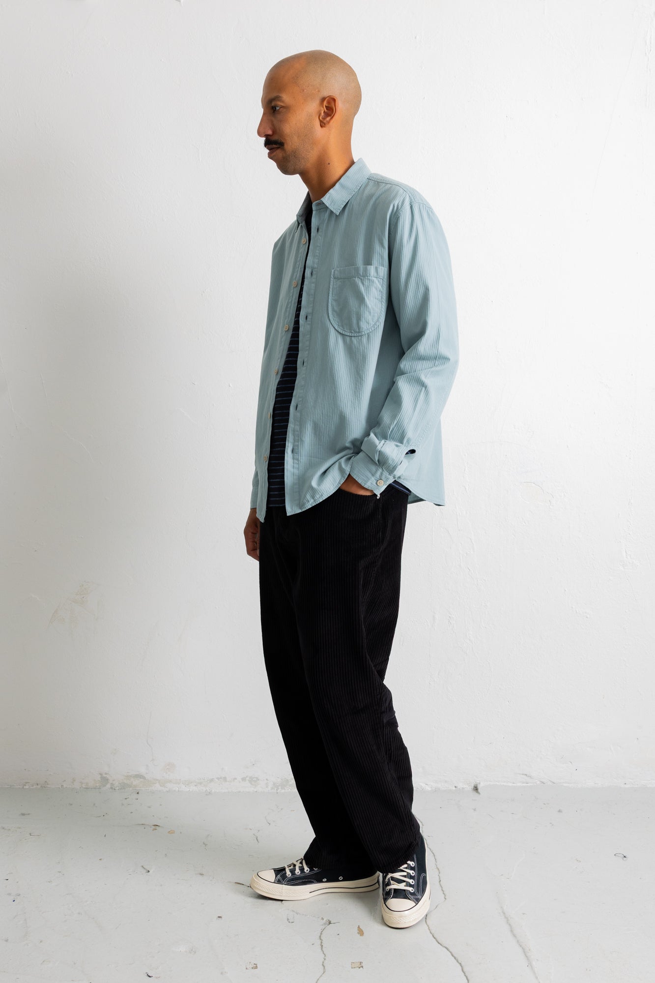 A man wearing a light blue long-sleeve button-down shirt, a black and white striped tie, black trousers, and black canvas sneakers, standing in profile against a white wall.