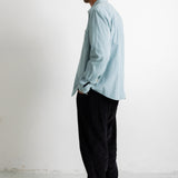 A man in profile wearing a light blue long-sleeve button-up shirt, black pants, and black canvas sneakers, standing against a plain white wall.