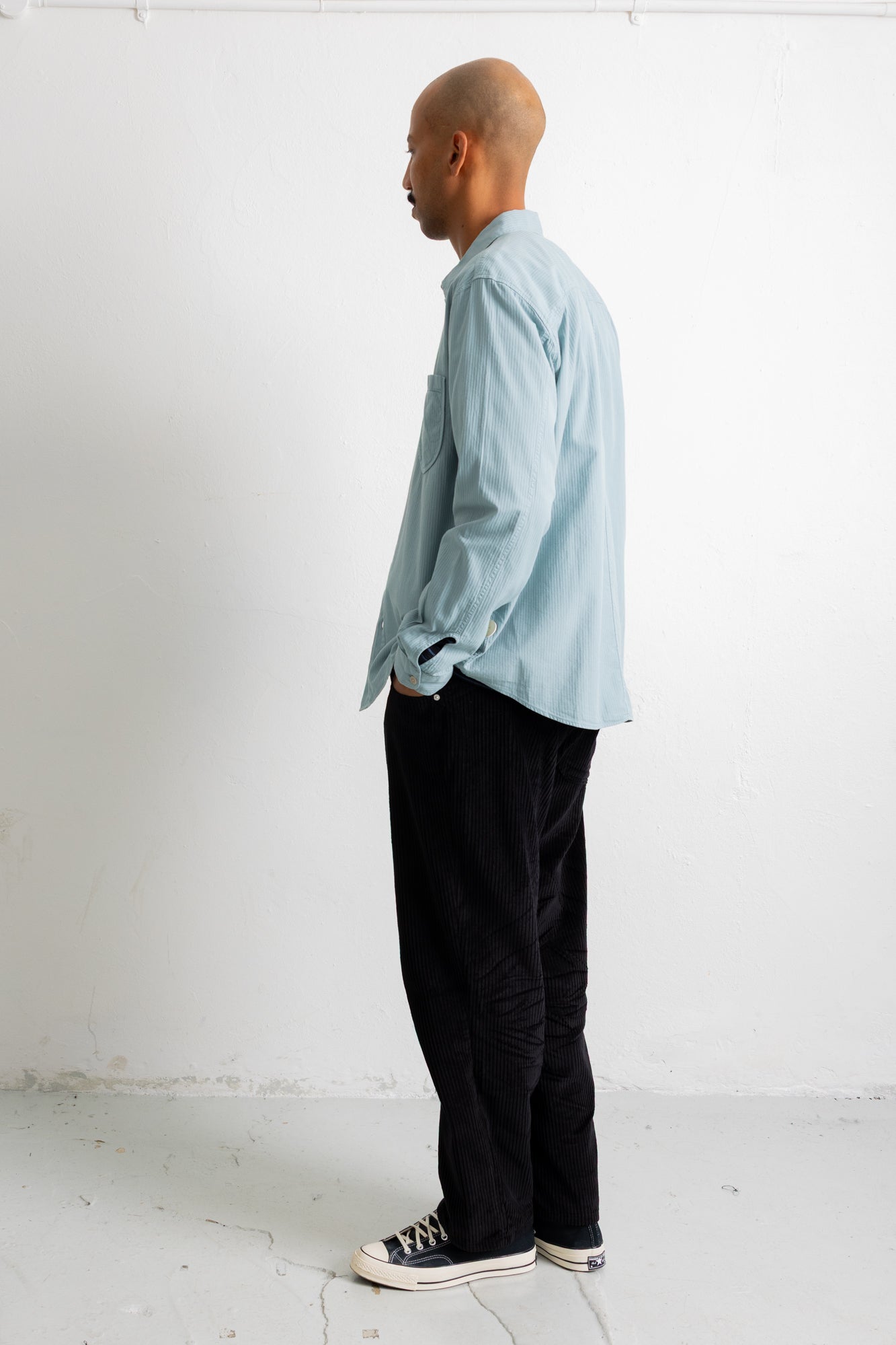 A man in profile wearing a light blue long-sleeve button-up shirt, black pants, and black canvas sneakers, standing against a plain white wall.