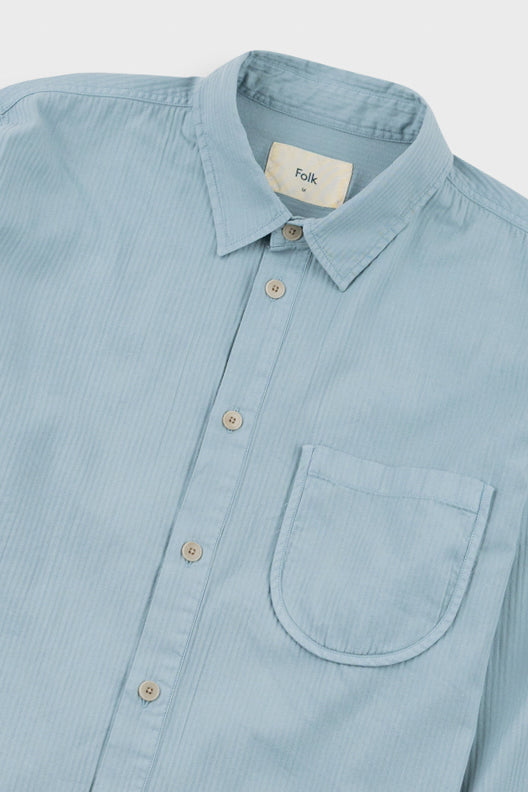 A close-up of a light blue button-down shirt with a collar, front buttons, and a chest pocket. The brand label inside the collar reads 'folk'.
