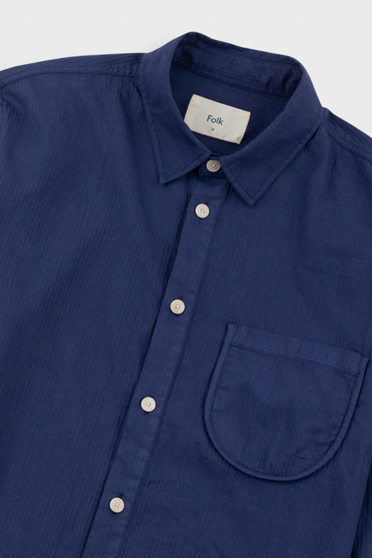 A close-up of a navy blue button-down shirt with a collar, white buttons, and a chest pocket. The brand label inside the collar reads 'Folk'.